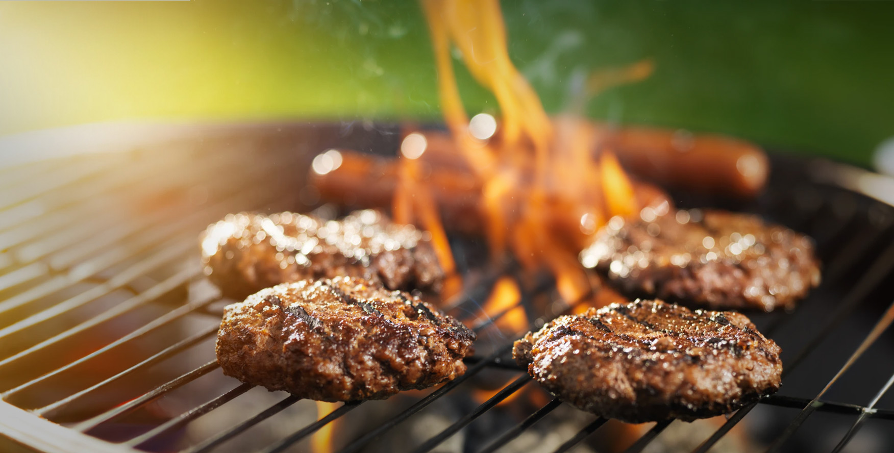 Beef burger patties and hot dogs cook over a flaming charcoal grill