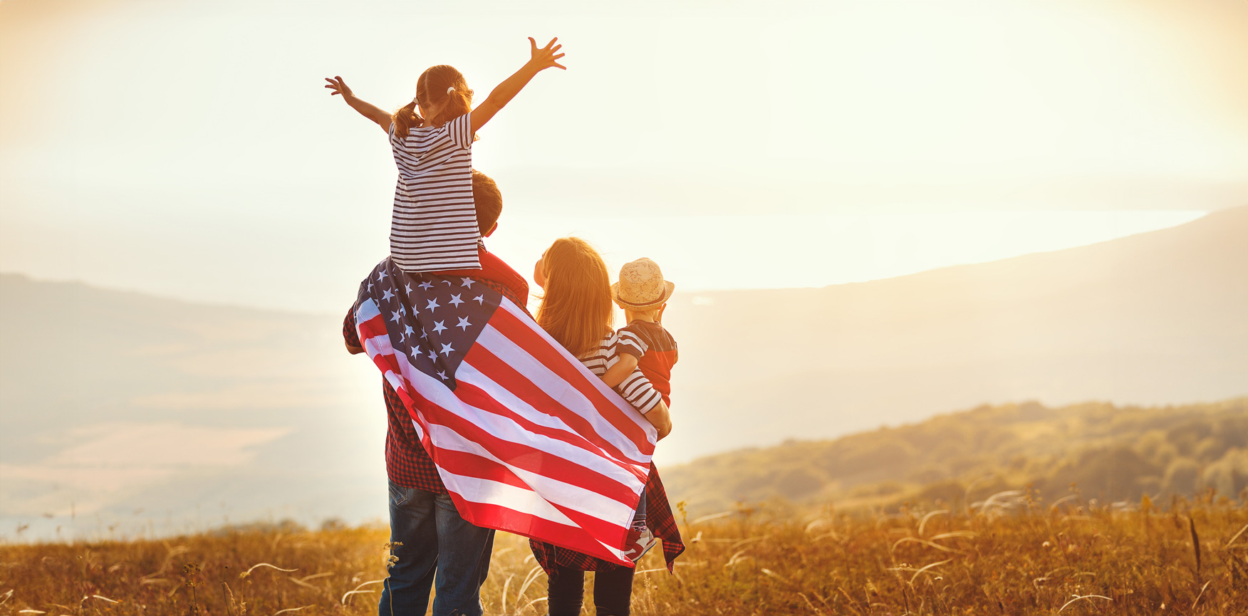 A father and mother hold their two children, wrapping the family in an American flag while looking over rolling hills
