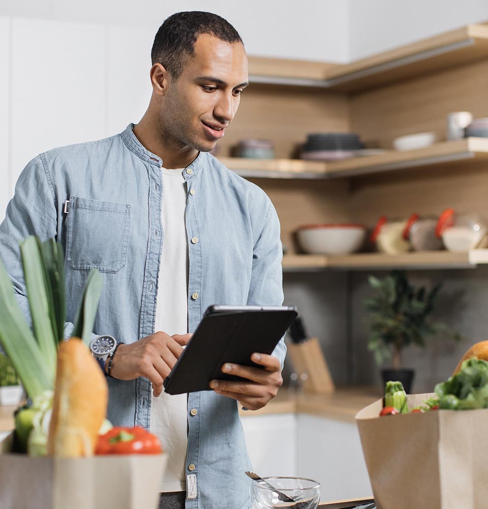 A smiling millennial in his kitchen, looking over his grocery purchases and checking them off the list on his tablet.