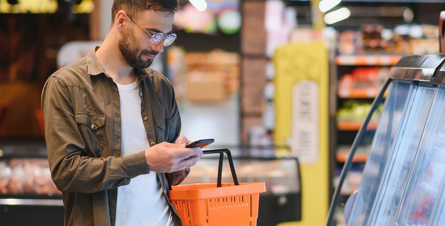 Man holding an orange shopping basket and looking at his phone at the meat case